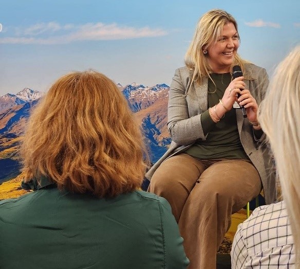 Joanna Costello, sitting outside in front of an audience holding a microphone, with mountains and blue sky as a backdrop.