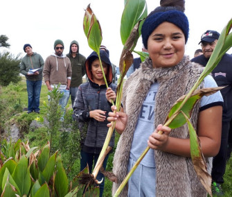 Photo: A group of Māori people stand on the edge of a river. A young girl is smiling and holding 2 tall green leaves in her hands.