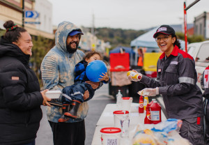 Photo: A man wearing a hoody and a cap carries a young child in his arm as he waits for a sausage. A Red Cross worker is smiling and hold mustard and a sausage.