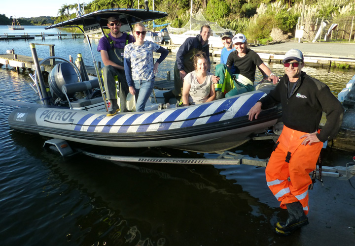 Photo: A patrol dinghy with 6 people in it, sitting on the edge of the water. One person in the foreground is leaning on the boat and smiling. He is wearing orange pants and a black shirt. 