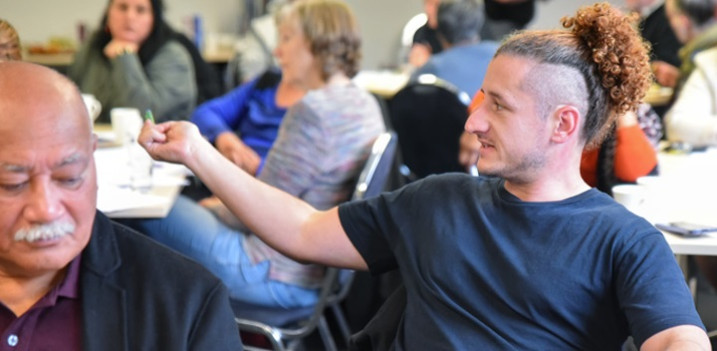 Photo: A man wearing a black shirt and curly hair tied up sits in a room of people at a desk. He is speaking a question and has his arm extended.