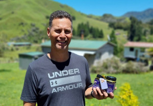 Photo: A smiling Māori man is standing on grass and holding 2 jars of honey. 