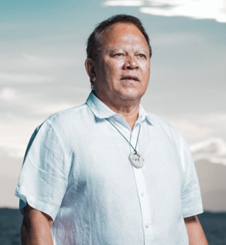 Photo: A Māori man stands next to the ocean. He wears a white shirt.