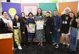 Photo: 9 people are standing and smiling in front of coloured boards in a room. The 2 women in the middle hold posters which say The Hive, We are the Future.