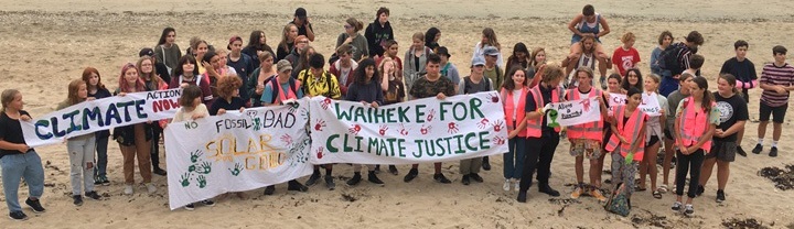 Photo: A large group of people stand on a beach holding signs. The large sign says Waiheke for Climate Justice.