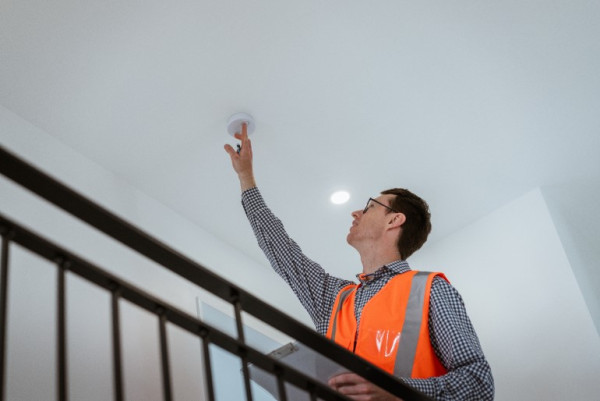 A person in an orange bib, holding a clip board, testing a domestic smoke alarm.