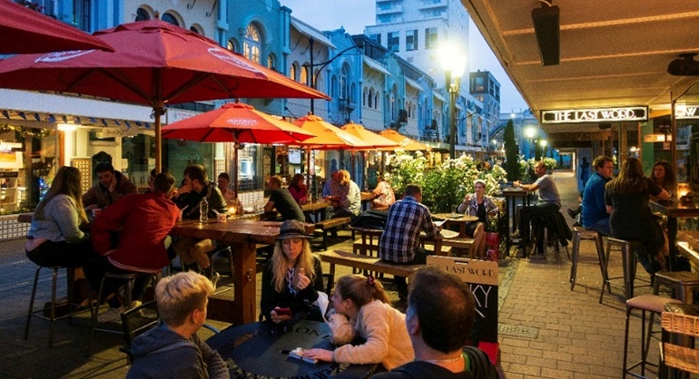 Patrons relax at tables in an outdoor cafe, on Christchurch's New Regent Street, sipping beverages and socializing in a vibrant, dusk atmosphere.