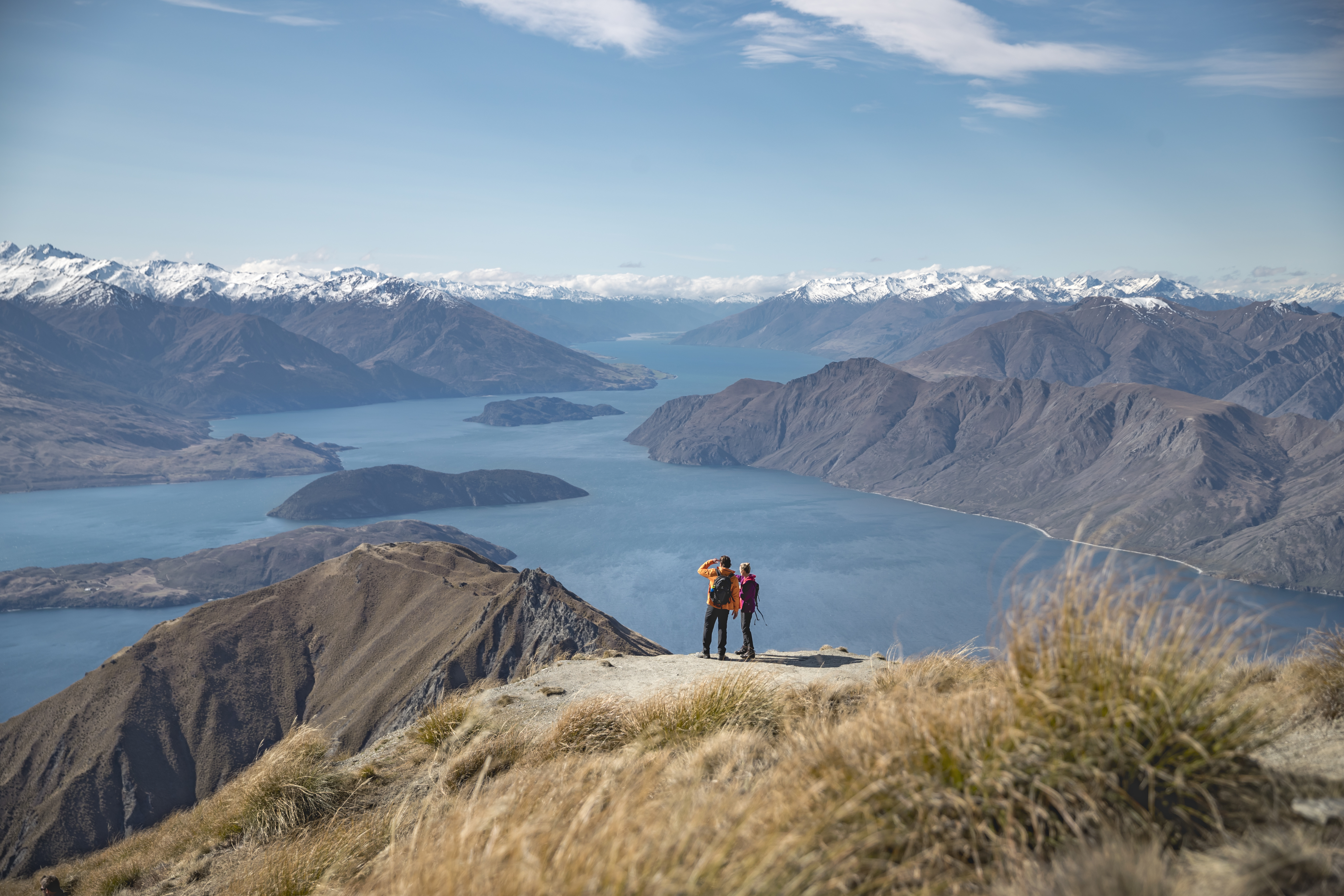 Two people standing on top of Roys Peak Wanaka looking out over the view of water and mountain ranges.