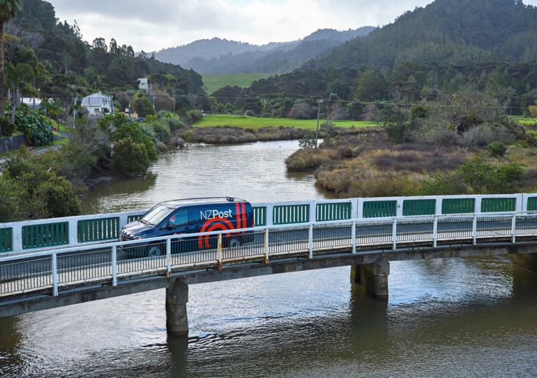 Aerial drone image of an NZ Post mail delivery van crossing a narrow bridge over a river. There are only a few houses shown, mainly landscape, implying that it may be a rural setting.