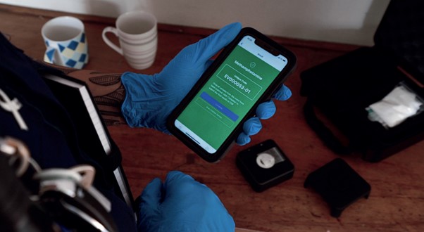 Photo of a Police officer in the field reviewing Lumi results on a smartphone. We can see their hands have rubber gloves on, there's a sample in the background, and the screens of a smartphone shows a positive result for methamphetamine.