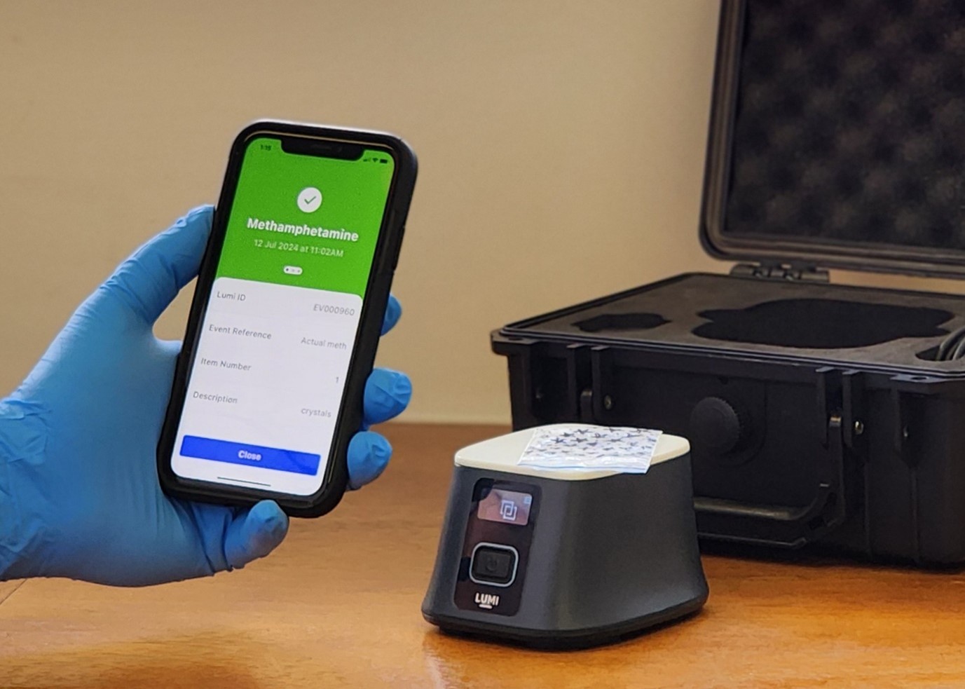 Officer in the field using a Lumi device and viewing results on a smartphone. We can see their hands have rubber gloves on, there's a sample in the background, and the screens of a smartphone shows a positive result for methamphetamine.