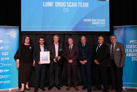 The Lumi™ Drug Scan team receiving a framed certificate at the 2023 Science New Zealand Awards. The photo shows 7 people standing together with event banners surrounding them.